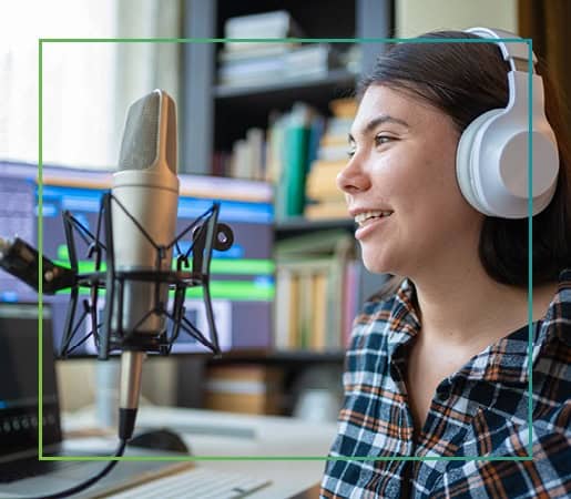 Woman wearing headphones speaking into a studio microphone at a desk, with a computer screen displaying audio recording software in the background.