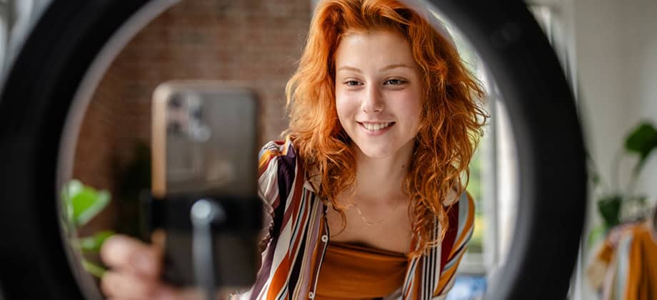 A person with red hair is adjusting the camera setup in front of a ring light
