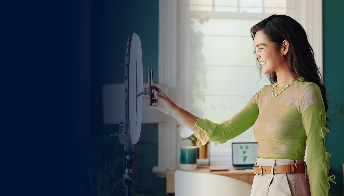 A woman stands indoors, smiling and adjusting a smartphone mounted on a ring light stand.