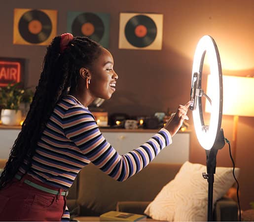 A woman adjusts a ring light in a cozy, decorated room with vinyl records on the wall and a lit lamp in the background.